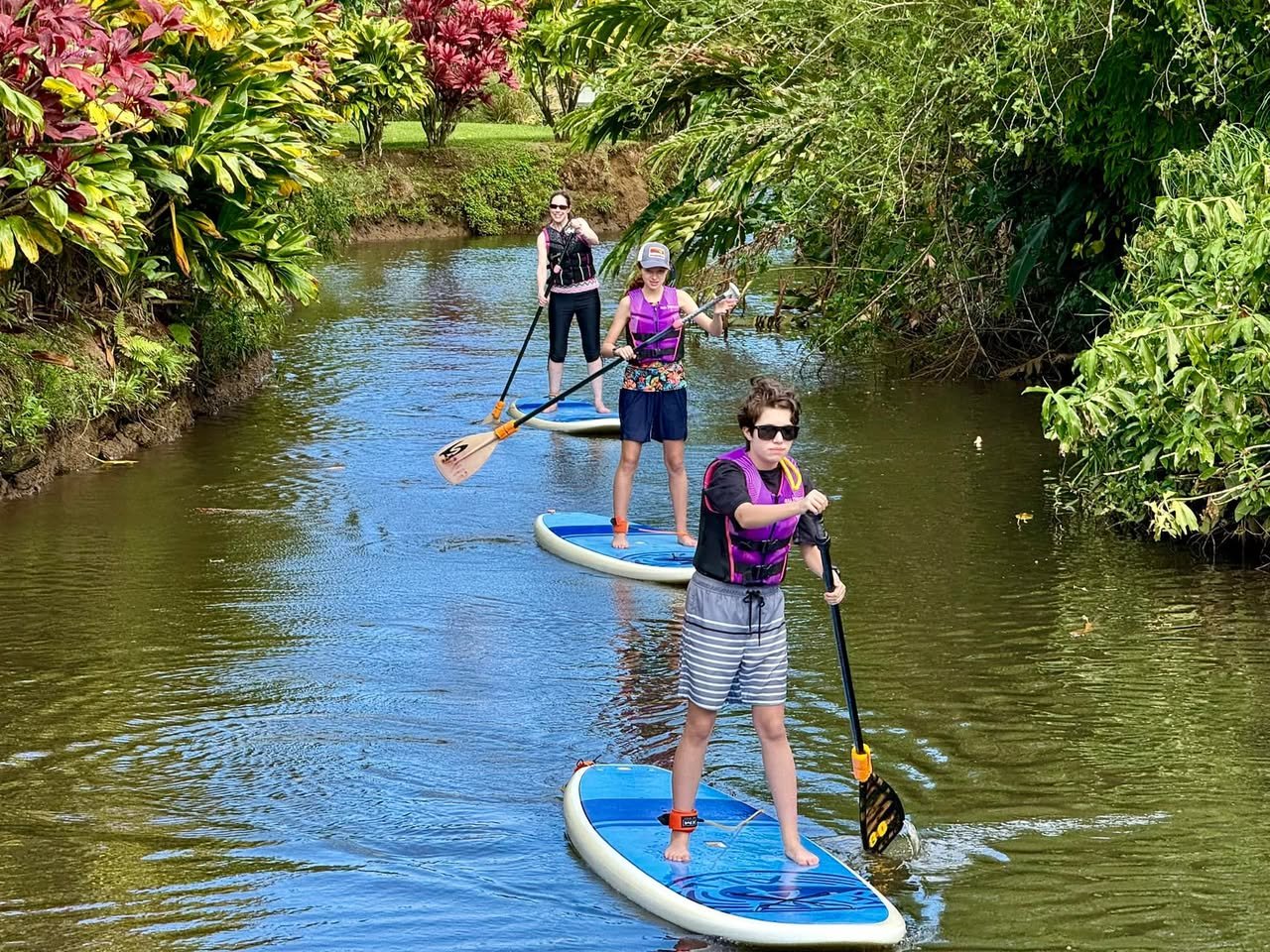 Three people paddleboard down a lush canal in vibrant life jackets, surrounded by tropical foliage. This serene scene captures a family-friendly outdoor adventure.