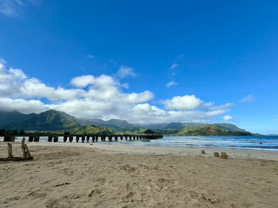 A panoramic view of Hana Bay featuring a sandy beach, a historic pier, and green mountains under a bright blue sky.