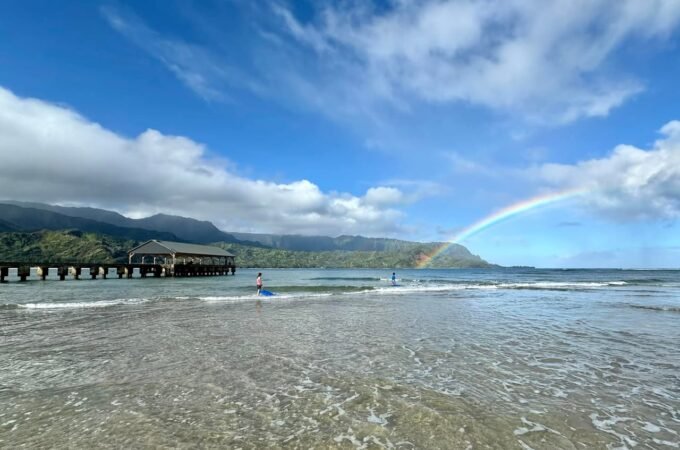 A rainbow arches over Crescent Bay Beach with a pier in the background and blue skies above.