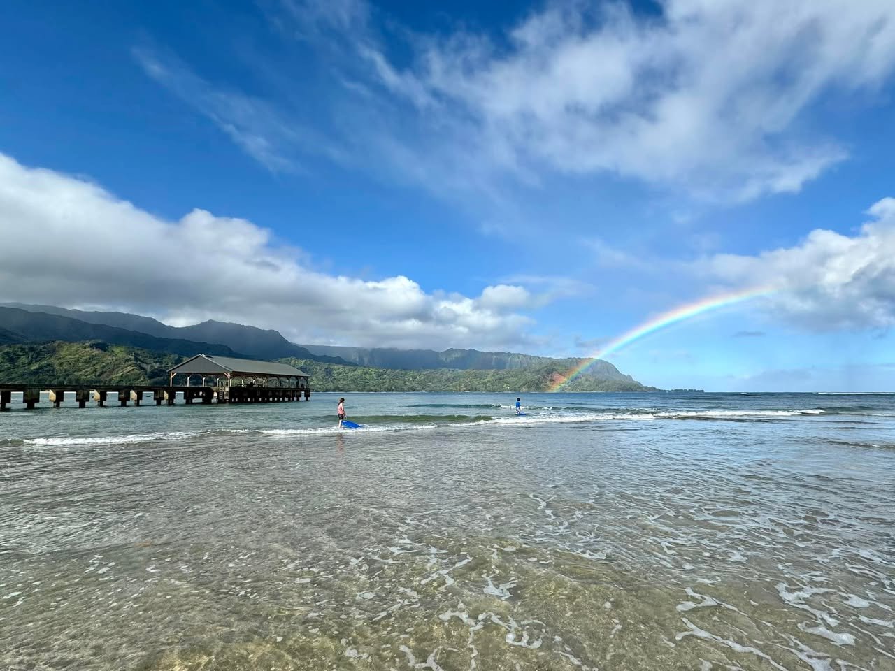Hanalei Bay, Kauai Hawaii A rainbow arches over Crescent Bay Beach with a pier in the background and blue skies above.