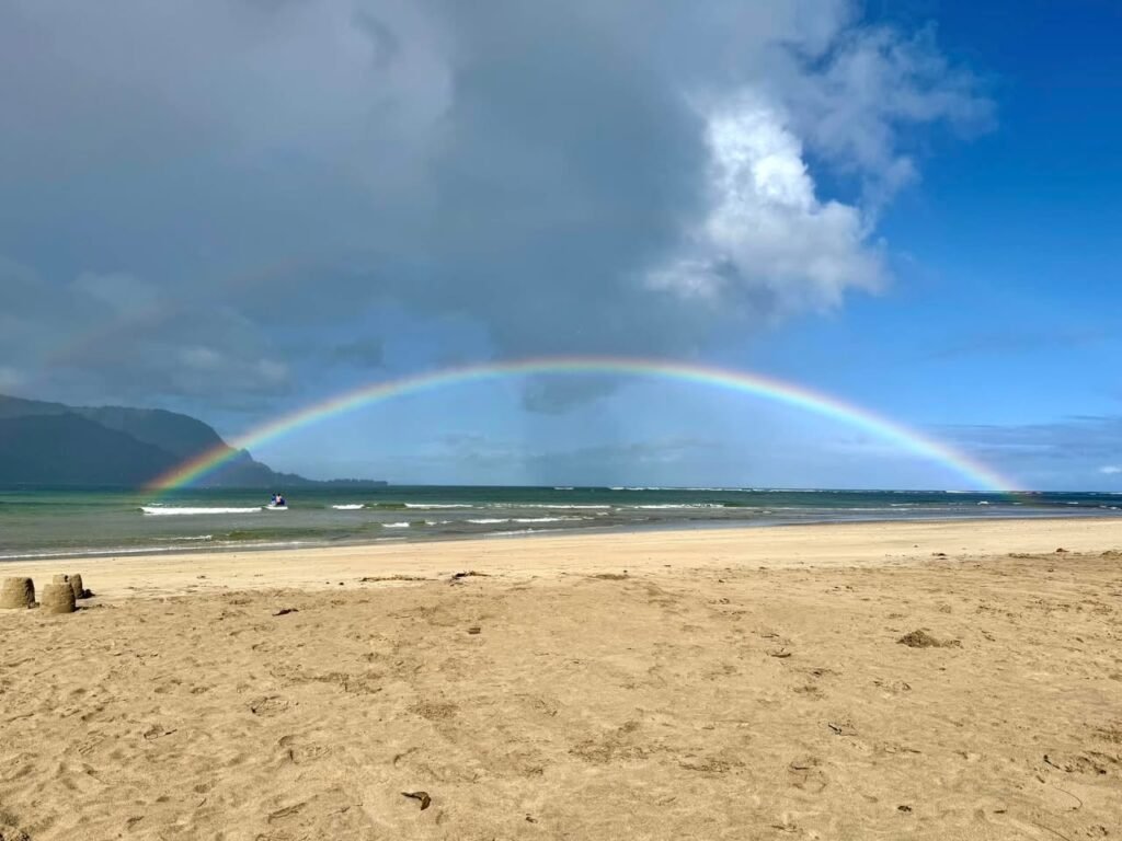A vibrant rainbow arches across a cloudy sky above a sandy beach. Gentle ocean waves roll onto the shore while the coastline stretches into the distance.