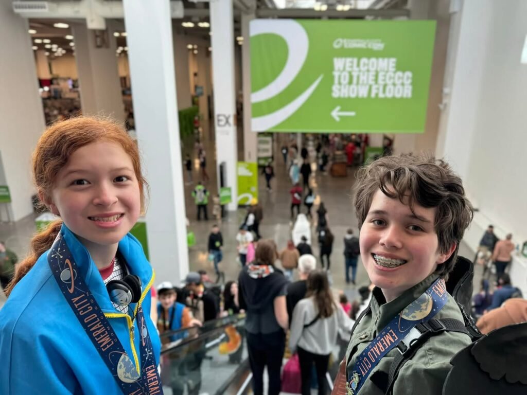 Two young attendees smile on a crowded convention show floor near a signage area.