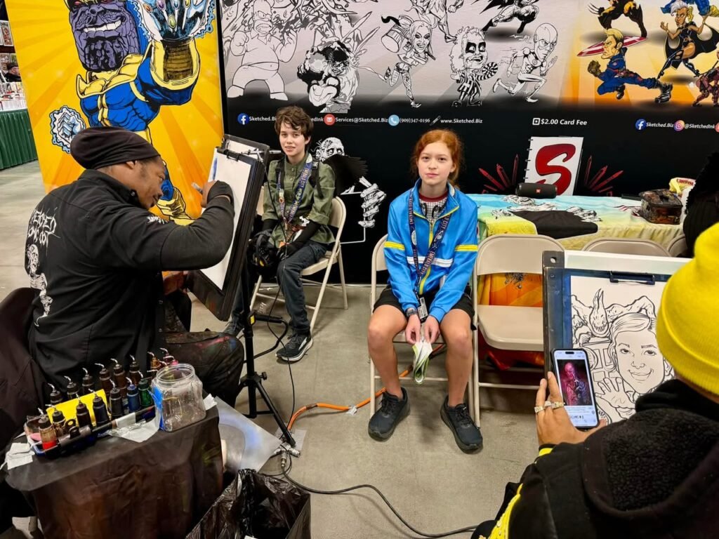 An artist works on a detailed illustration at a convention booth, with attendees and a superhero backdrop.