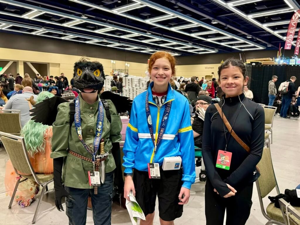 Three friends posing together at a comic-con convention, including someone wearing a detailed bird costume in a busy hall.