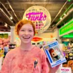 A traveler smiles while holding a Funko Pop box in a brightly lit retail store. The iconic Funko globe sign hangs prominently for visitors to see.