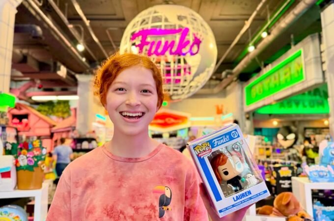 A traveler smiles while holding a Funko Pop box in a brightly lit retail store. The iconic Funko globe sign hangs prominently for visitors to see.