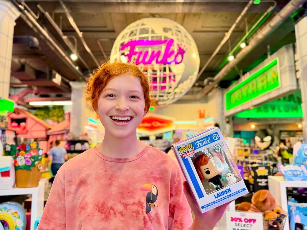 Funko HQ A traveler smiles while holding a Funko Pop box in a brightly lit retail store. The iconic Funko globe sign hangs prominently for visitors to see.