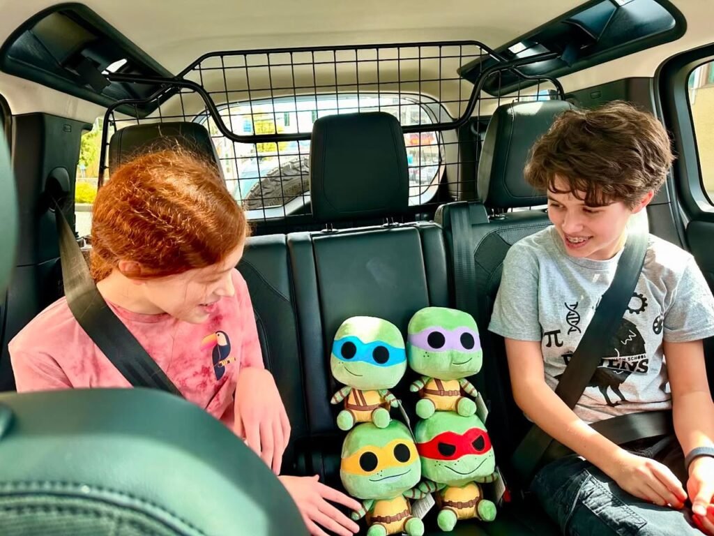 Two people sitting in a car's back seat beside four Teenage Mutant Ninja Turtles plush toys.