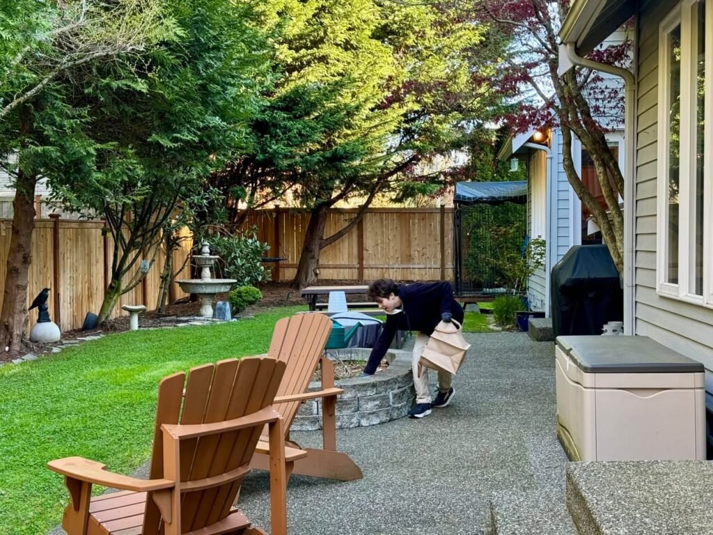 A cozy backyard patio featuring an Adirondack chair and stone fire pit. The lush green lawn and mature trees create a peaceful atmosphere.