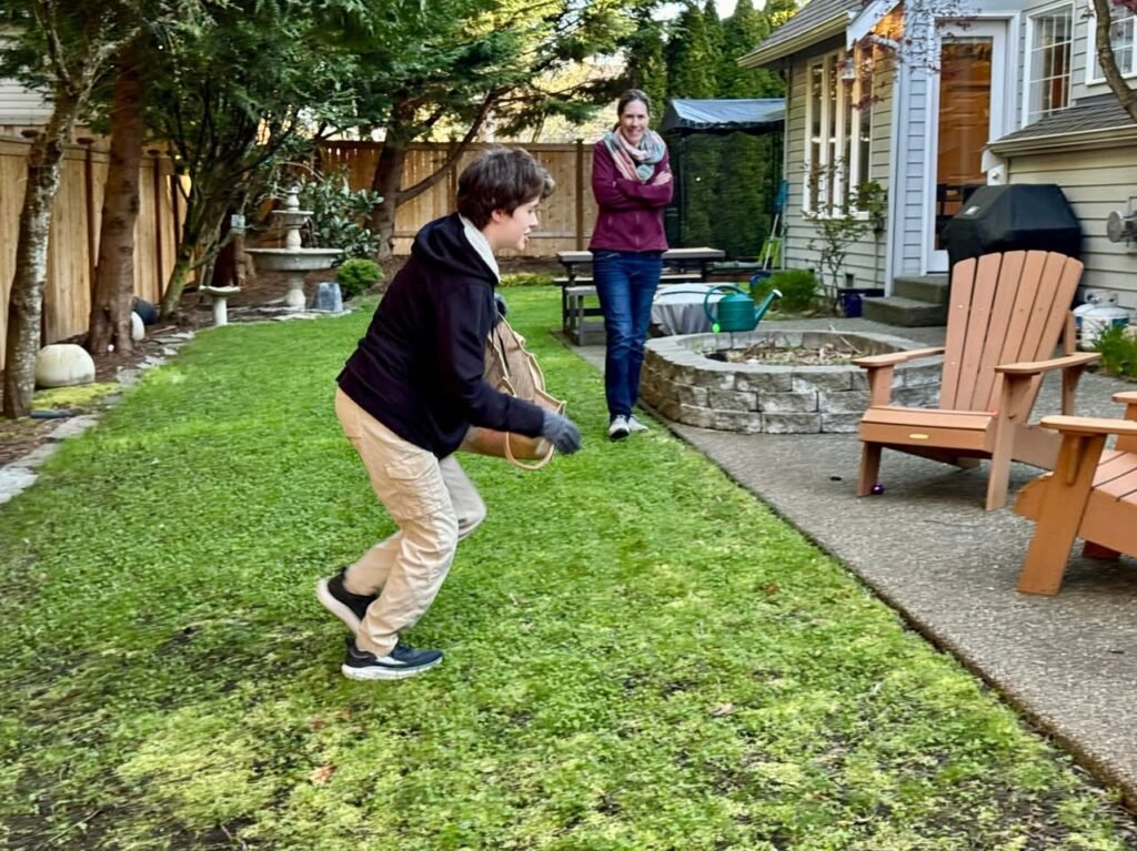 Two people playing ball in a suburban backyard with a stone fire pit and Adirondack chairs.