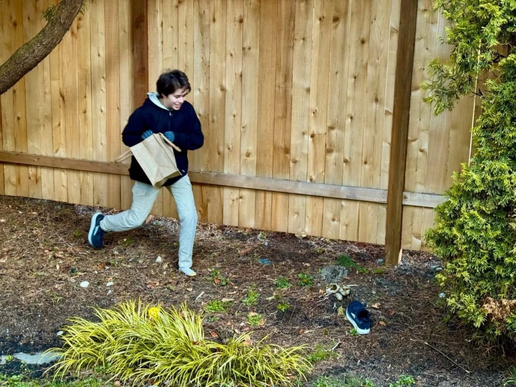 A child in dark clothing and light pants runs through a muddy backyard along a wooden fence, holding a brown paper bag. Green shrubs and grass border the dirt path.