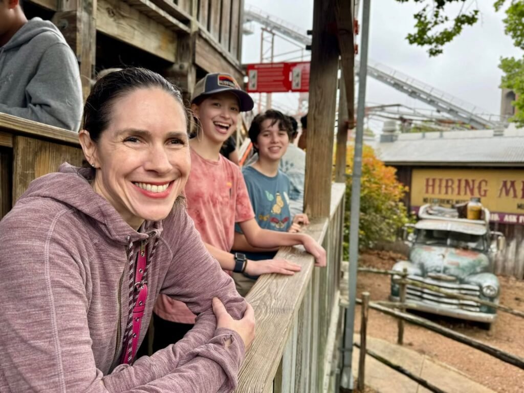 A family enjoys a day at an amusement park, watching the rides and attractions from a wooden stand.