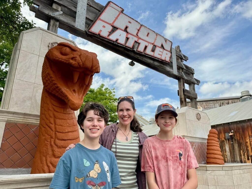 Smiling family posing in front of the Iron Rattler roller coaster sign on a sunny day.