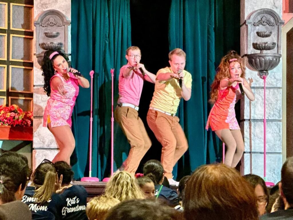 Young performers sing energetically on a stage with green curtains and pink microphone stands.