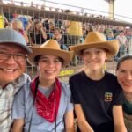 Six Flags Fiesta Texas A happy family wearing cowboy hats enjoys a rodeo event from the spectator stands. They are smiling for a photo in front of the metal enclosure.