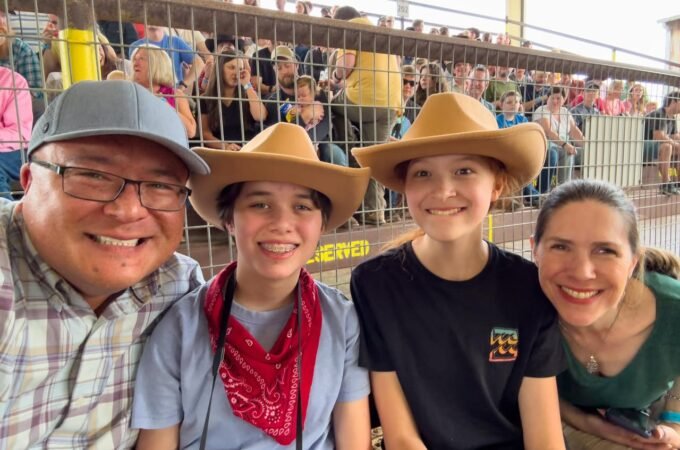 A happy family wearing cowboy hats enjoys a rodeo event from the spectator stands. They are smiling for a photo in front of the metal enclosure.