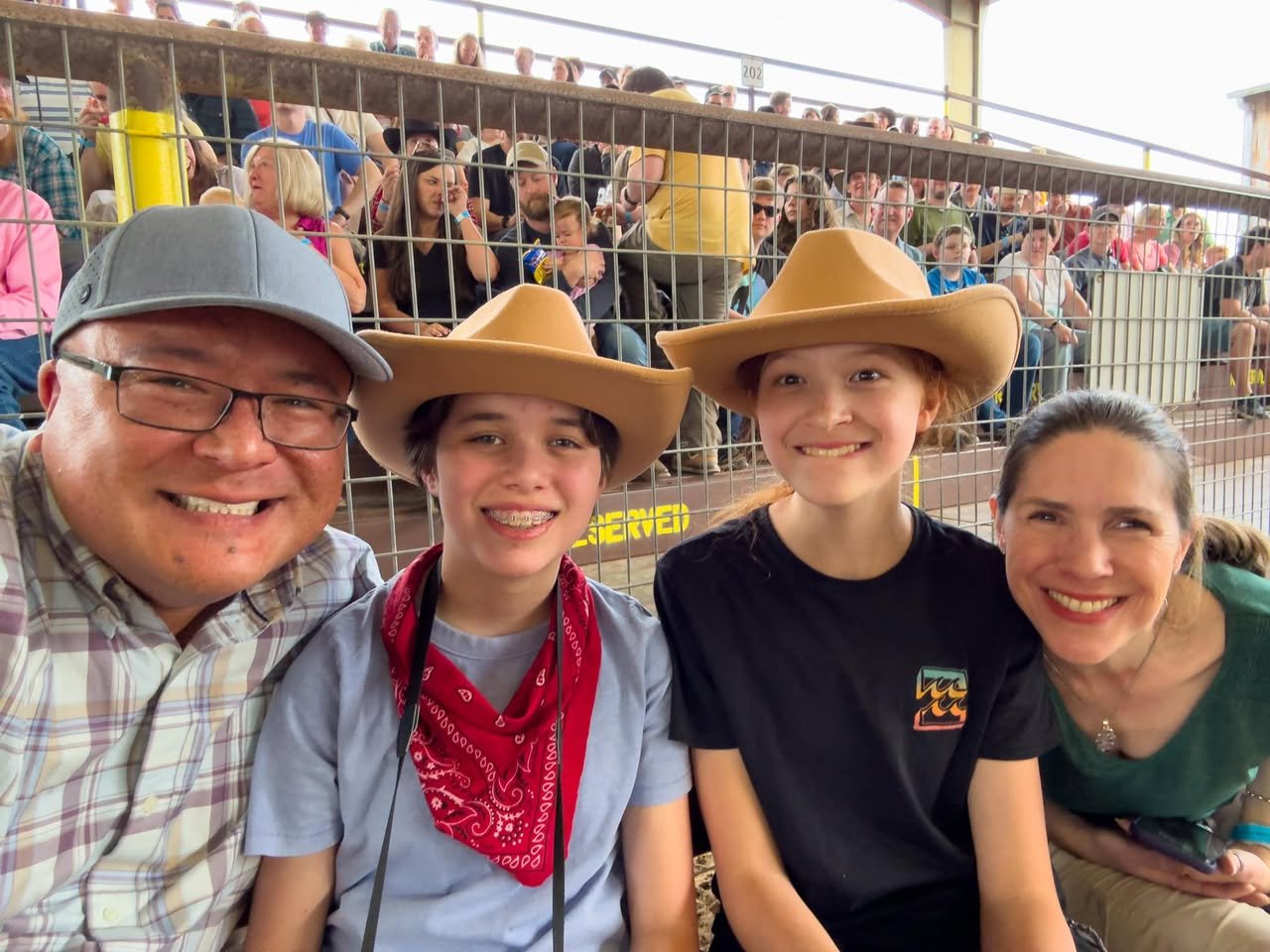 Our First Rodeo A happy family wearing cowboy hats enjoys a rodeo event from the spectator stands. They are smiling for a photo in front of the metal enclosure.