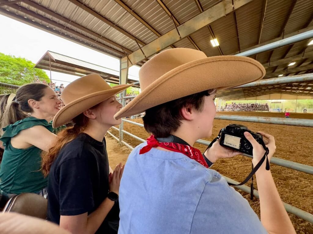 A group of people wearing cowboy hats at a rodeo event, one holding a camera. The image shows a covered rodeo arena with metal fencing and a dirt surface.