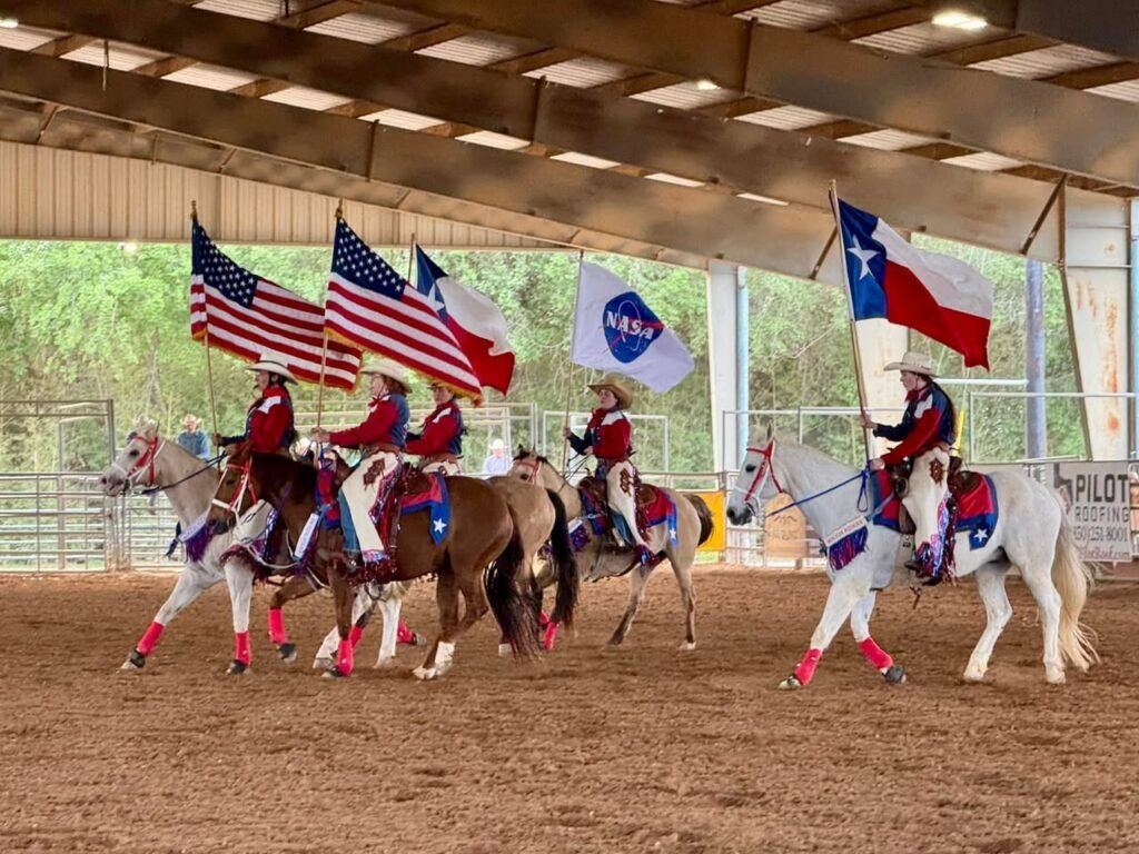 Riders on horseback carrying American, Texas, and NASA flags during a show at an indoor arena.