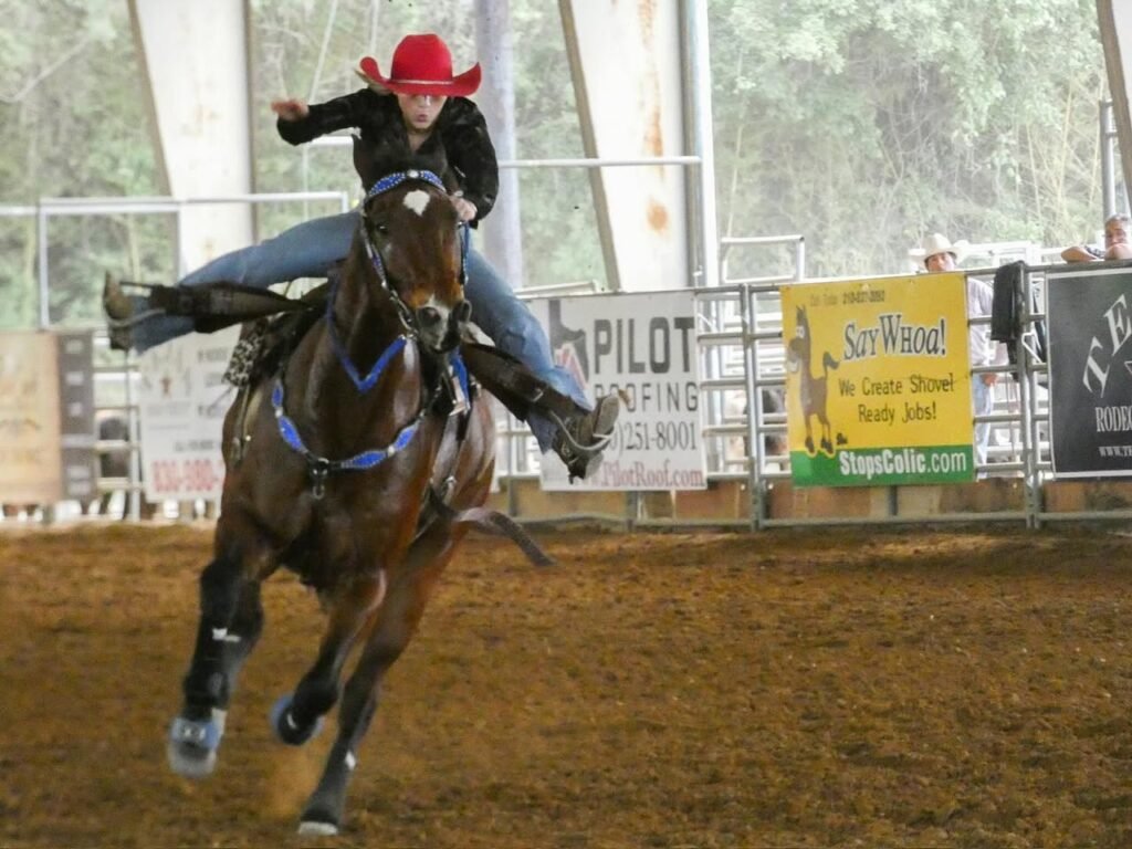 A rider in a red hat on a bucking horse at an indoor rodeo event. The horse is mid-buck in a dirt arena surrounded by advertising banners.
