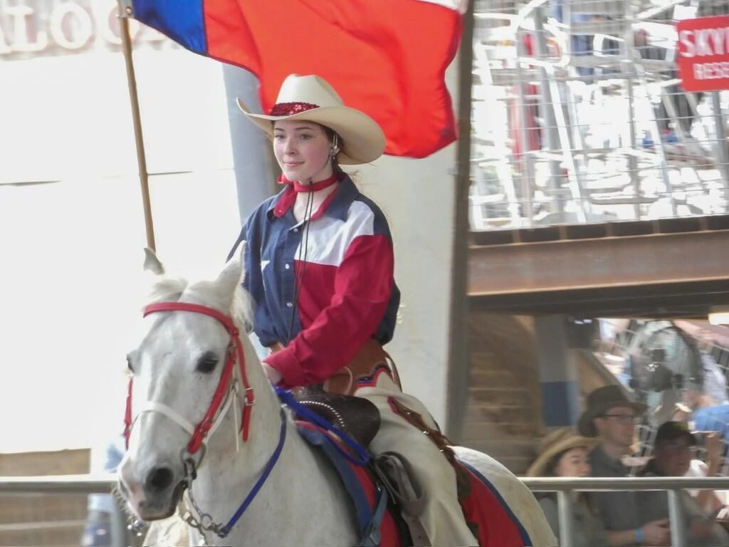 A person in a cowboy hat rides a white horse while holding a red and blue flag. The rider wears a colorful plaid jacket in a parade setting.