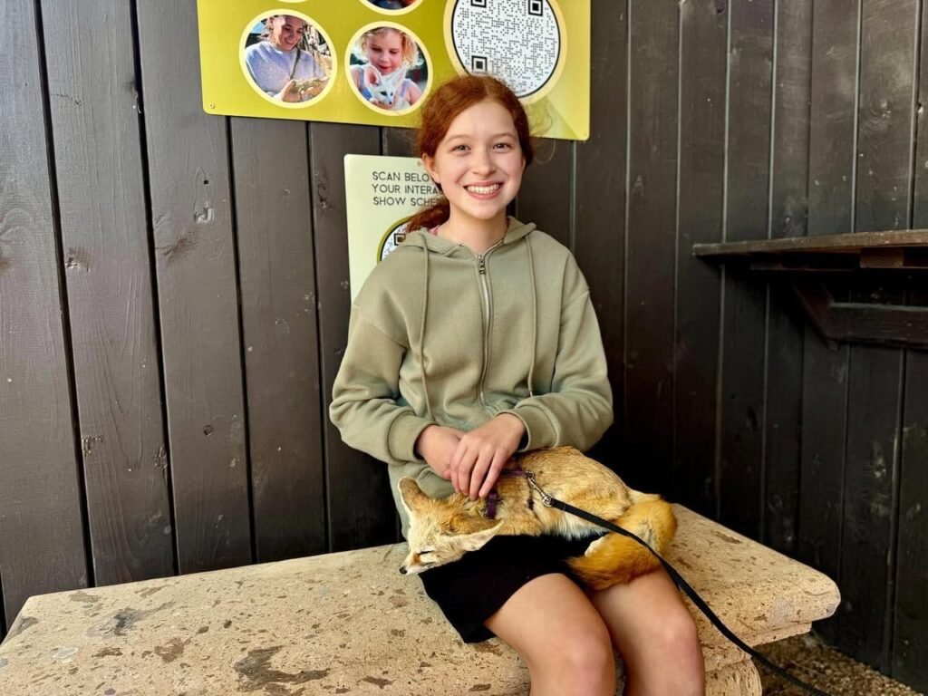 Two girls smiling with a small animal on a stone surface, with a colorful poster on a wooden wall.