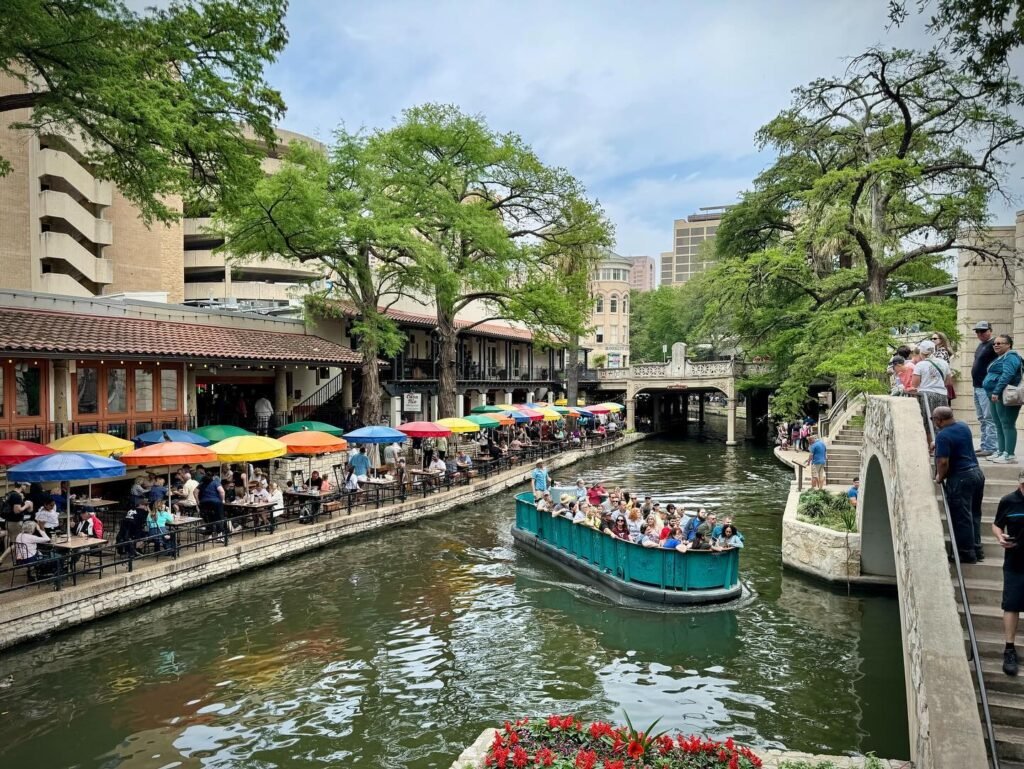 A boat filled with tourists glides down a canal lined with colorful umbrellas and people dining along the water's edge in a city setting. The scene includes historic buildings and a stone bridge.