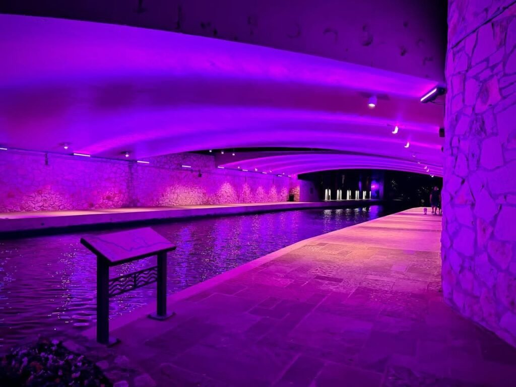 A vibrant purple-lit underground canal walkway within the ancient Roman catacombs, featuring stone arches, reflective water, and a small bench.
