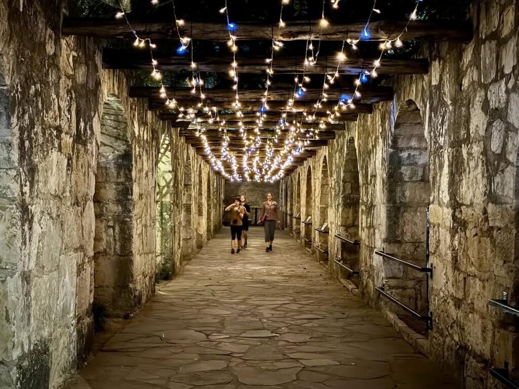 People wander a stone-paved alleyway with old walls under a canopy of multicolored string lights.