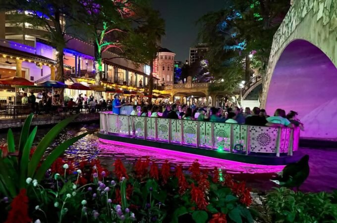 A brightly lit boat with a pink underglow travels along a river at night, passing under a purple-lit arch bridge. Colorful marinaside buildings and vibrant red flowers line the canal.
