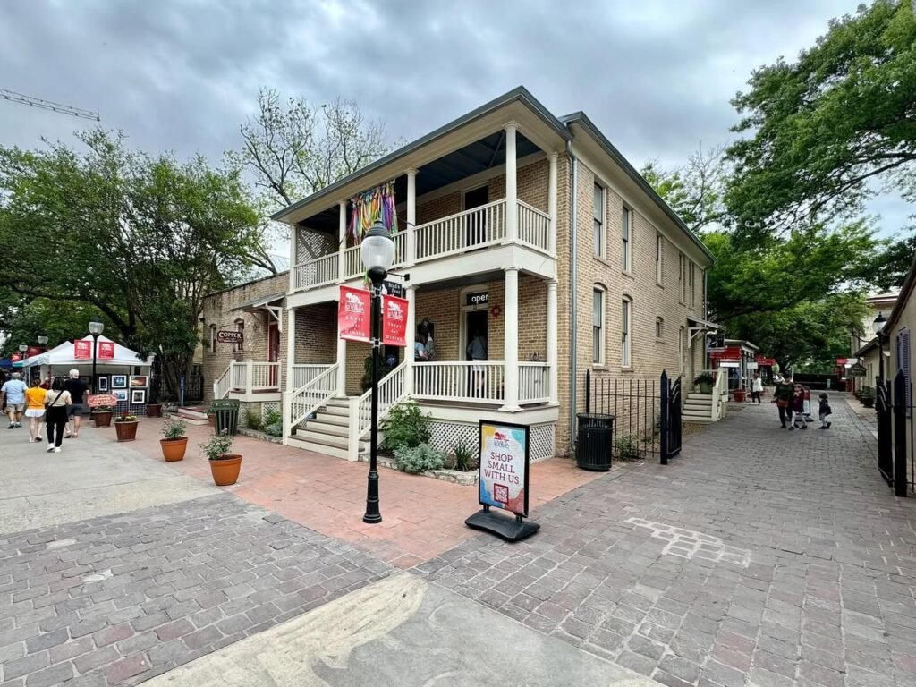 A street-level view of a historic two-story building with balconies adjacent to a paved pedestrian plaza featuring potted plants and promotional signage.