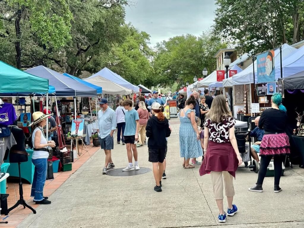 Attendees browse colorful vendor booths under a canopy of trees at an outdoor art market.