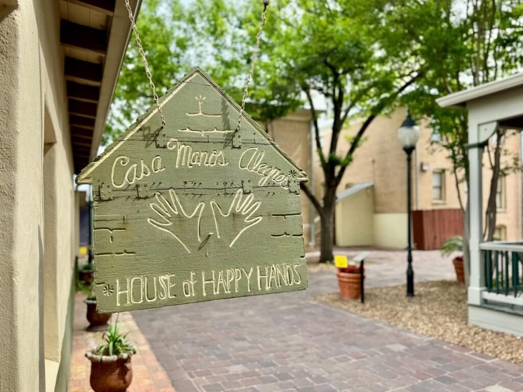A whimsical hanging sign illustration of a house with open hands and cheerful text marks the entrance to Casa Manos Alegres in a courtyard.