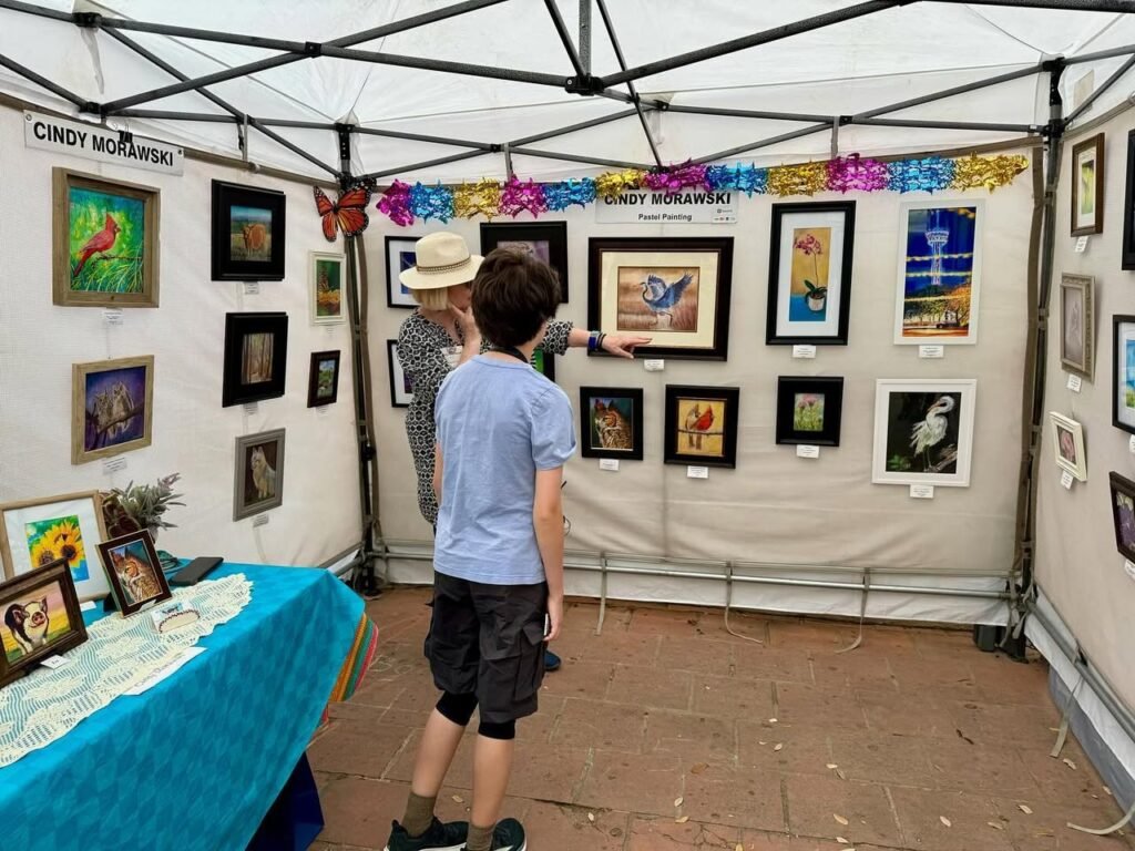A person explores various framed artworks on a display wall inside a festival tent with a table of crafts nearby.