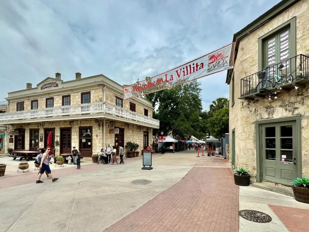 A view of the shops and walkways at La Villita in San Antonio, Texas. The image displays a banner for La Villita hanging between stone buildings.