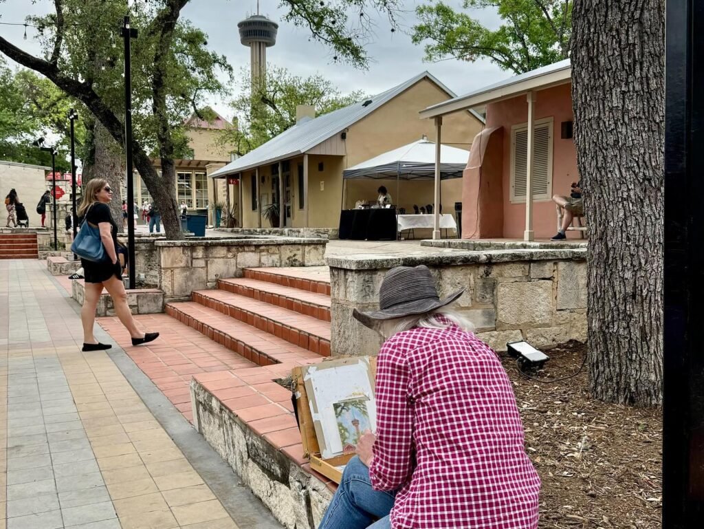 An artist in a plaid shirt and hat works on a canvas on a public walkway. A person walks past the artist near a small plaza with steps and trees.