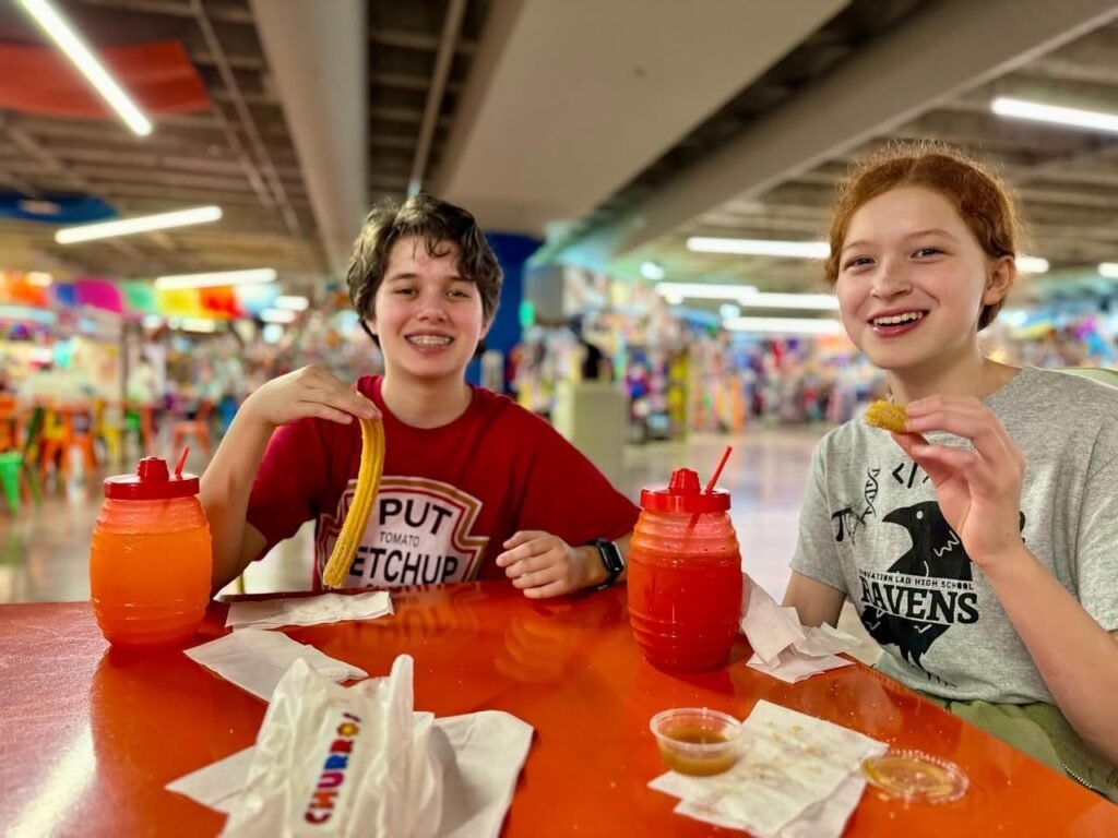 Two people sitting at a red table in a carnival setting with colorful condiment dispensers. A Chick-fil-A bag and small food items are visible on the table.