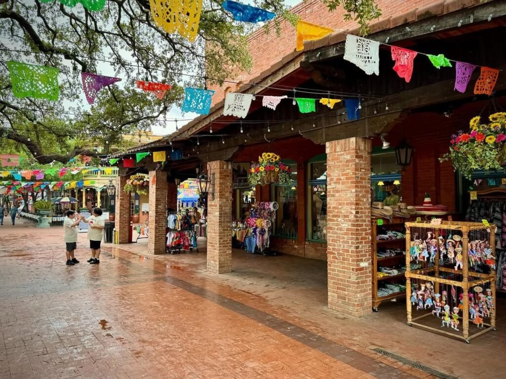 Colorful flags and banners adorn a rainy outdoor market with brick storefronts and a wet cobblestone walkway.