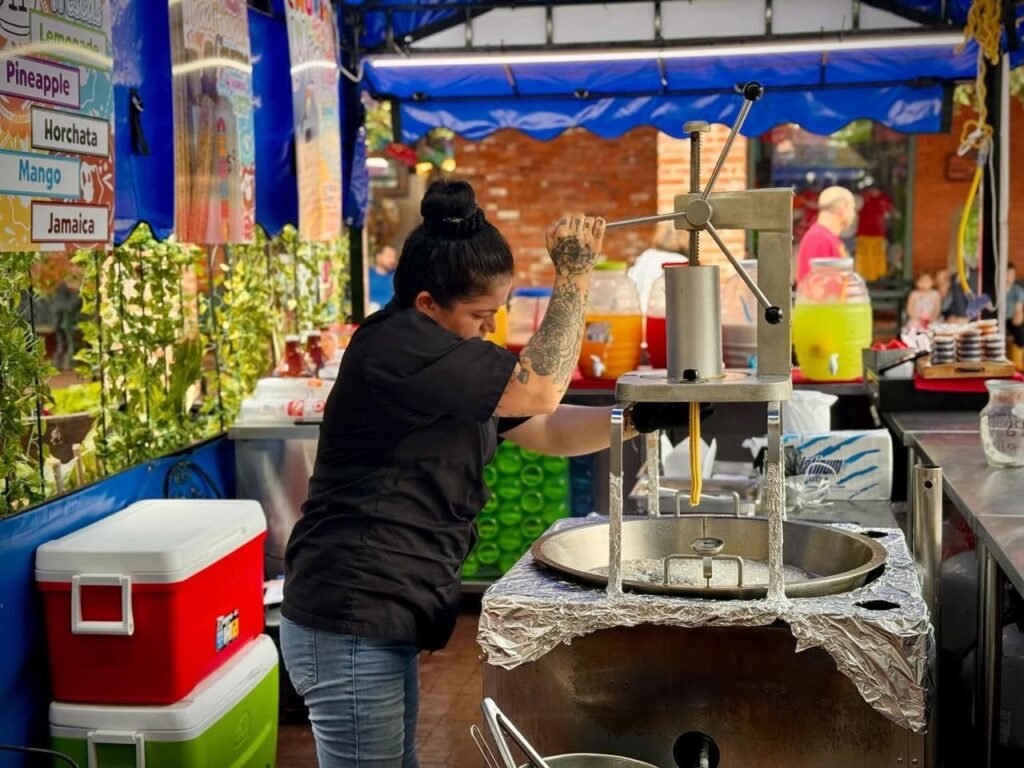 A person operates a large manual juicer at an outdoor market stall, surrounded by colorful coolers and potted plants.