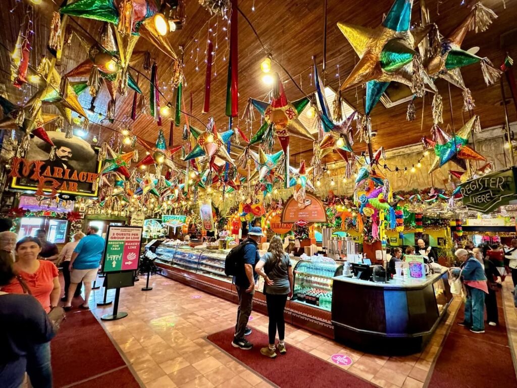 A vibrant indoor market with colorful star decorations, customers browsing at the counter, and a lively shopping atmosphere.