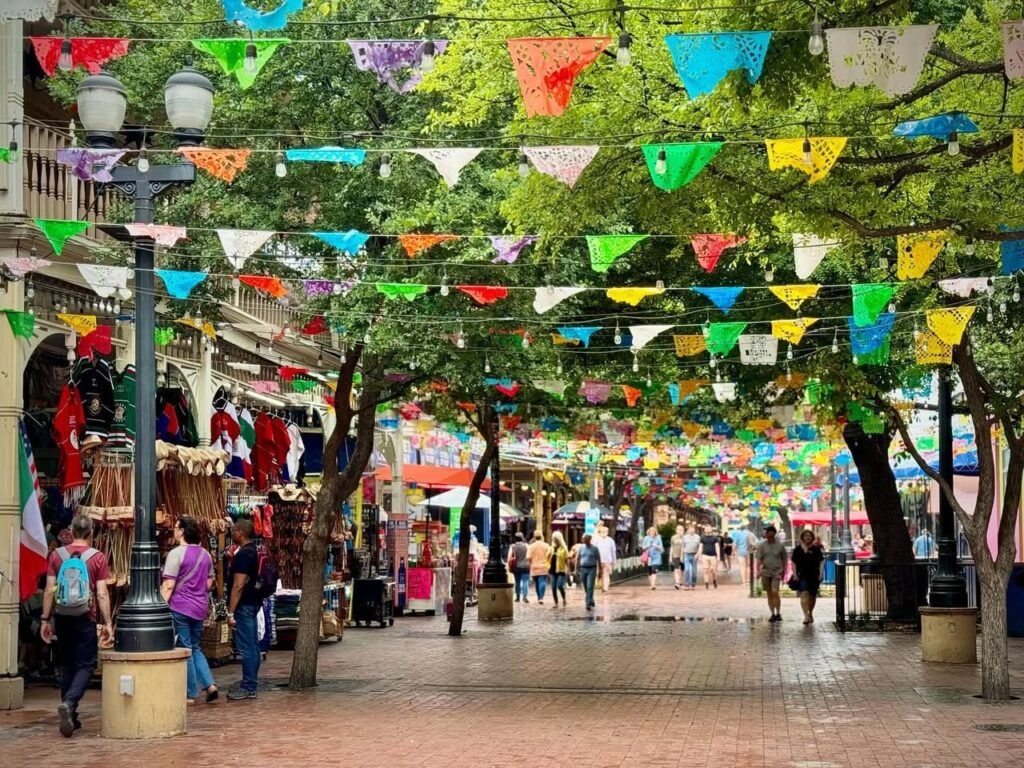 Vibrant bunting hangs above a bustling outdoor market street with visitors and green trees