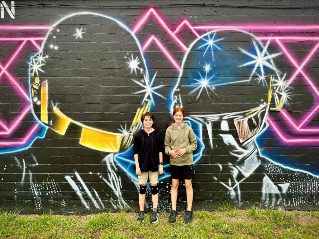 Two people pose in front of a black graffiti artwork featuring pink and blue neon geometric shapes.