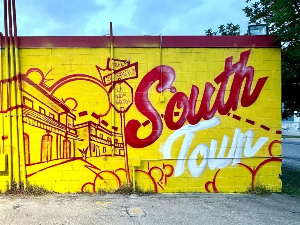 A vibrant yellow and red city wall mural featuring the words 'South Town' in bold cursive. Old-fashioned buildings decorate the mural against a bright yellow background.