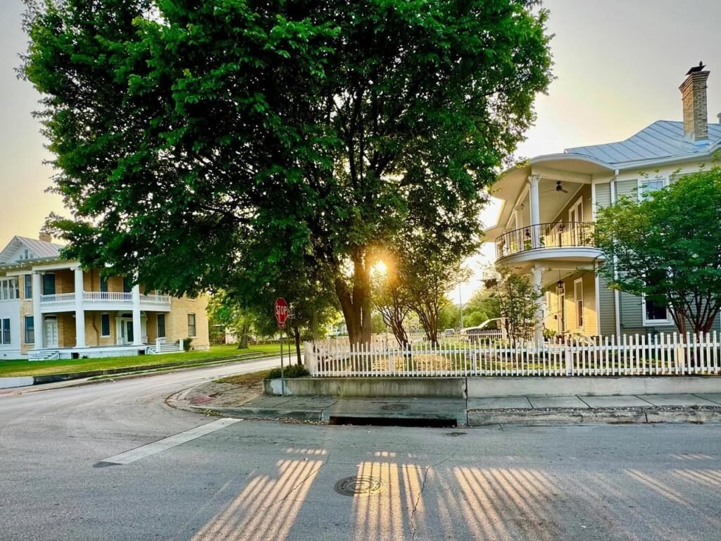 A historical street scene featuring an ornate yellow mansion with white verandas, surrounded by lush green trees.