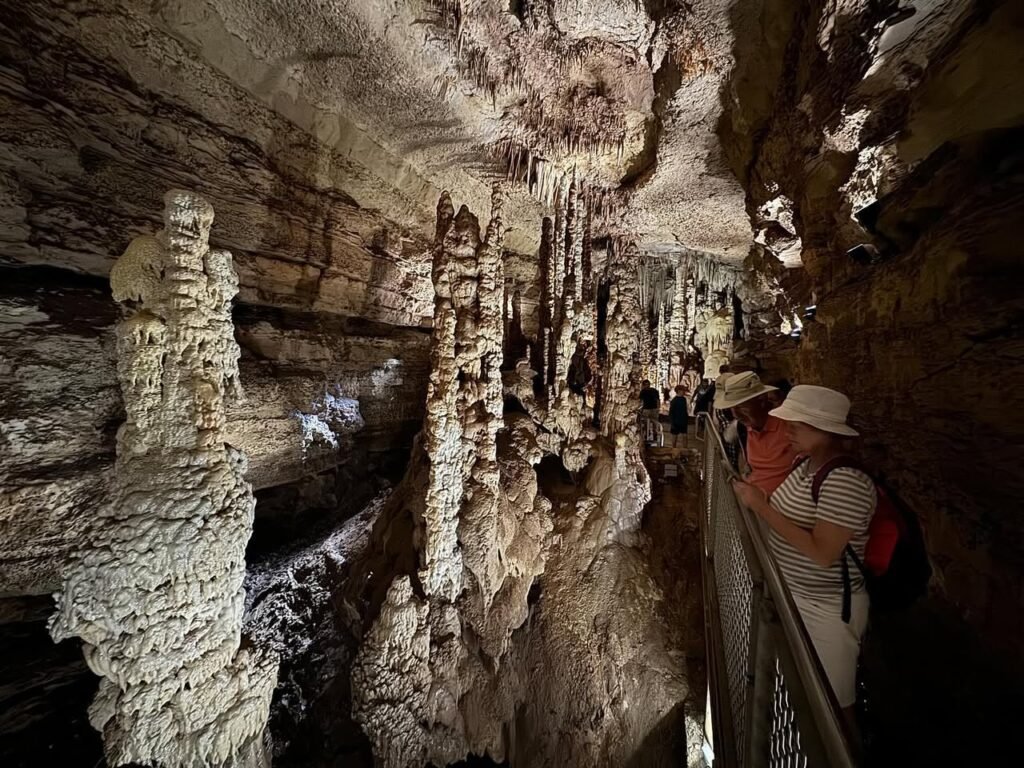 Visitors explore a cave featuring prominent stalactites and stalagmites under artificial light.