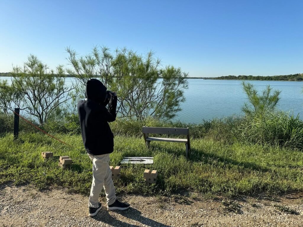 A person takes lake photography while standing on a gravel path. A wooden bench and calm waters complete this scenic view.