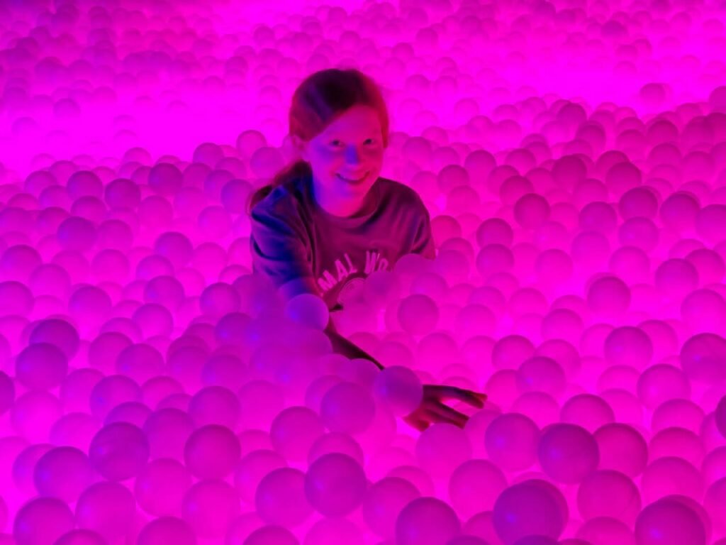 A person smiles while submerging in a sea of bright pink plastic balls illuminated by vibrant lighting.