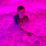 A person smiles while submerging in a sea of bright pink plastic balls illuminated by vibrant lighting.