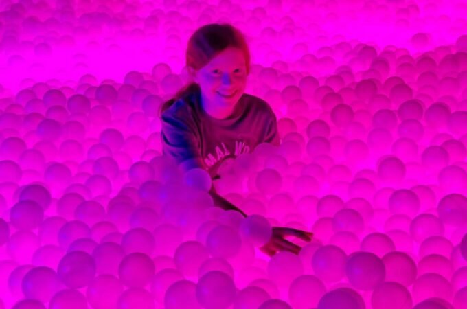 A person smiles while submerging in a sea of bright pink plastic balls illuminated by vibrant lighting.