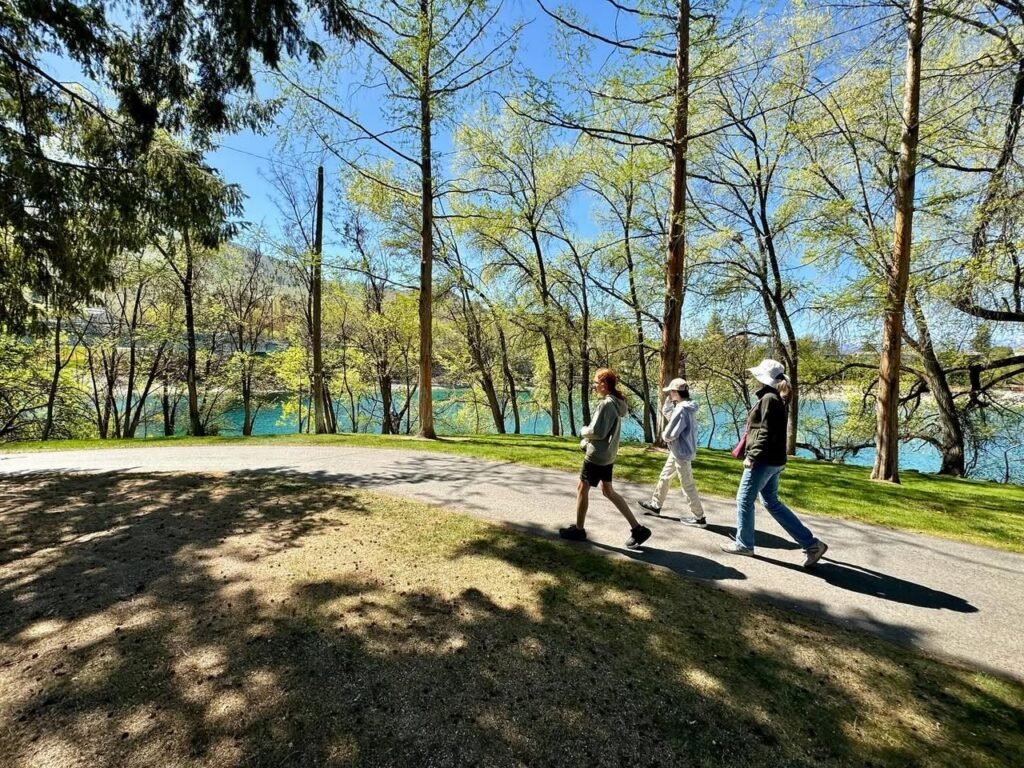 People walking on a shaded paved path beside a vibrant blue-green lake surrounded by spring foliage under a clear sky.